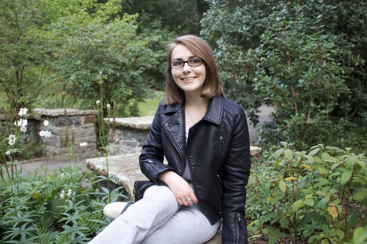 Elin sitting on a stone wall that zig zags through green plants and trees. Her right arm rests on her leg and she's smiling at the camera