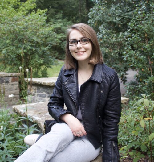 Elin sitting on a stone wall that zig zags through green plants and trees. Her right arm rests on her leg and she's smiling at the camera