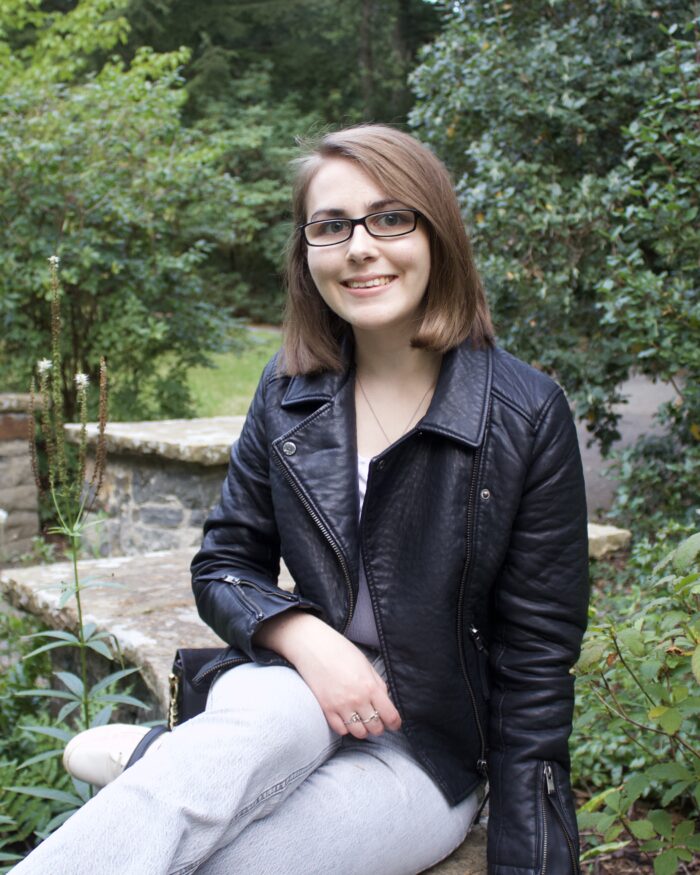 Elin sitting on a stone wall that zig zags through green plants and trees. Her right arm rests on her leg and she's smiling at the camera