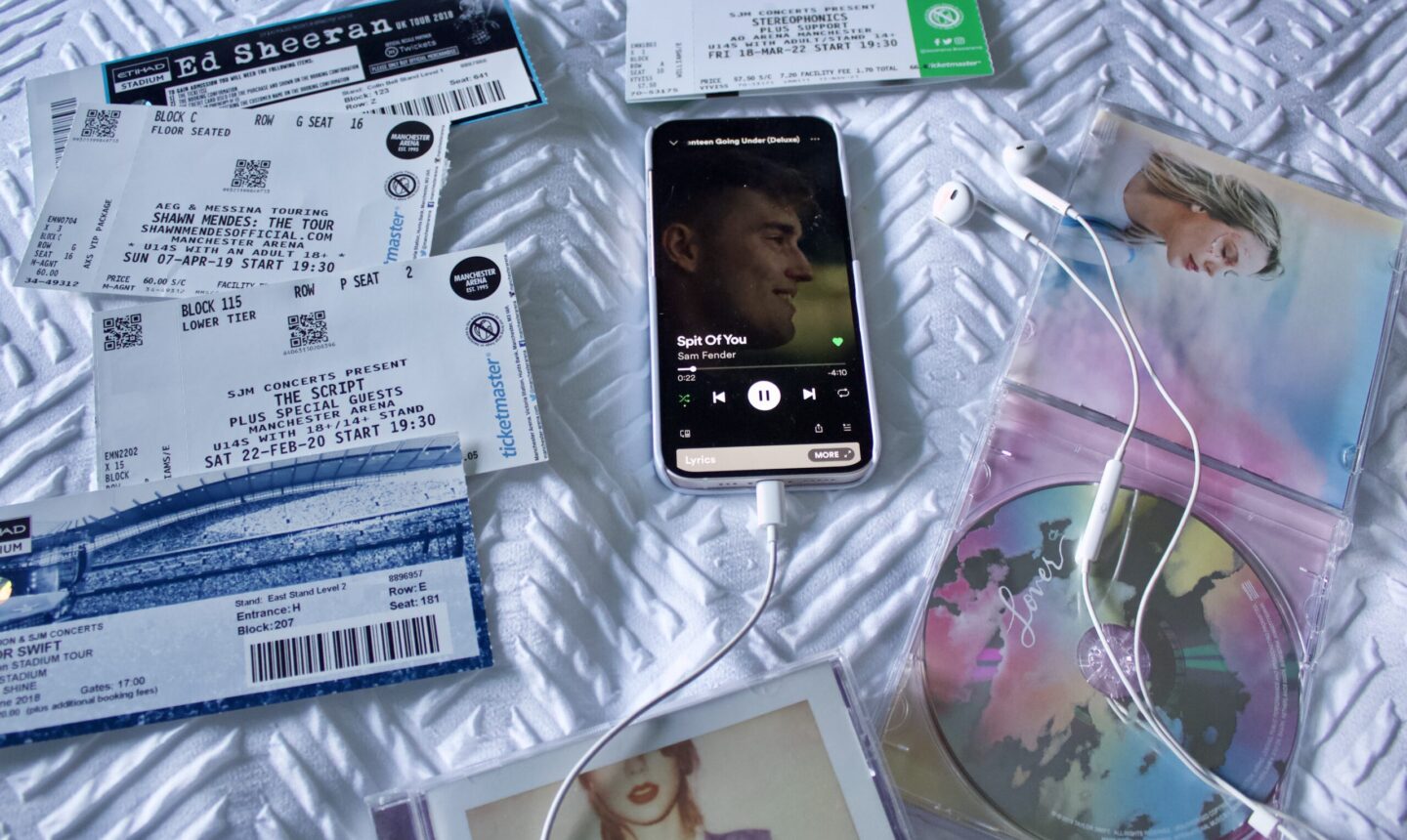 The song Spit of You by Sam Fender plays on an iPhone which has earphones plugged into it. The phone is surrounded by a collection of concert tickets including Ed Sheeran, The Script and Stereophonics. There are two Taylor Swift albums in the corner, one is open showing the CD inside