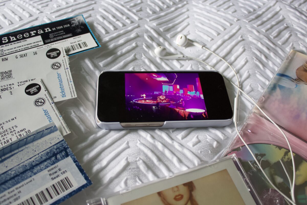 An iPhone showing a photo of The Script on a stage which is lit up in bright pink, blue and purple. The phone is surrounded by a collection of concert tickets which include Stereophonics, Ed Sheeran and Taylor Swift. Two Taylor Swift albums rest beside the phone and there is a pair of earphones on top of them