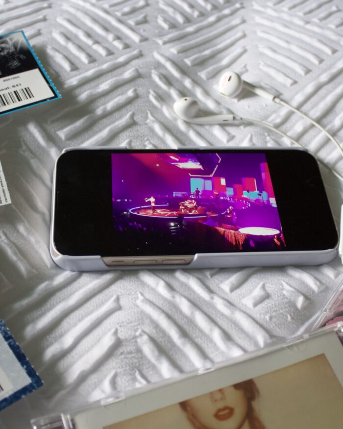 An iPhone showing a photo of The Script on a stage which is lit up in bright pink, blue and purple. The phone is surrounded by a collection of concert tickets which include Stereophonics, Ed Sheeran and Taylor Swift. Two Taylor Swift albums rest beside the phone and there is a pair of earphones on top of them