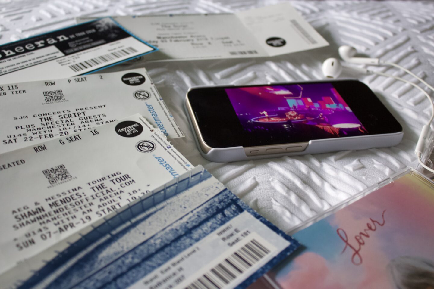 An iPhone showing a photo of The Script on a stage which is lit up in bright pink, blue and purple. The phone is surrounded by a collection of concert tickets which include Stereophonics, Ed Sheeran and Taylor Swift. Two Taylor Swift albums rest beside the phone and there is a pair of earphones on top of them