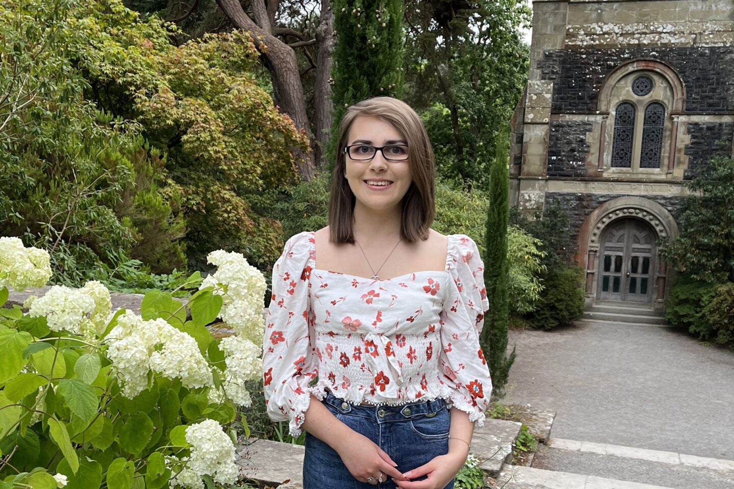 Elin standing on top of steps which lead down to a small stone building with wooden doors and windows. The building is flanked by trees. She wears a white and red floral printed top which has slightly puff sleeves which fall to just below the elbow. It's paired with dark blue high waisted jeans, a silver heart shaped necklace and silver rings. Her shoulder length brown hair is worn straight and she's smiling at the camera.