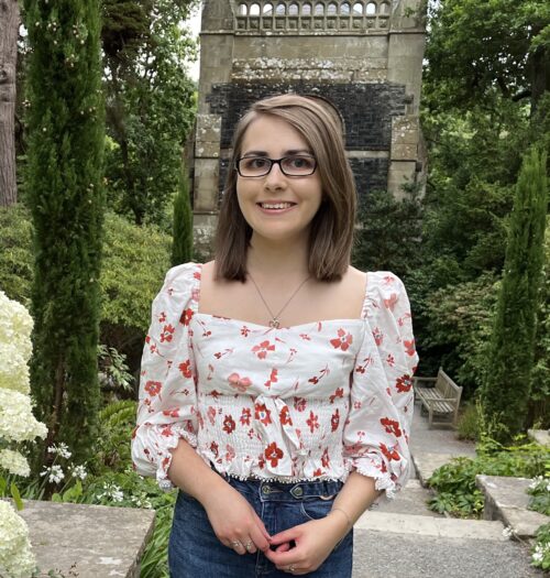 Elin standing on top of steps which lead down to a small stone building with wooden doors and windows. The building is flanked by trees. She wears a white and red floral printed top which has slightly puff sleeves which fall to just below the elbow. It's paired with dark blue high waisted jeans, a silver heart shaped necklace and silver rings. Her shoulder length brown hair is worn straight and she's smiling at the camera.