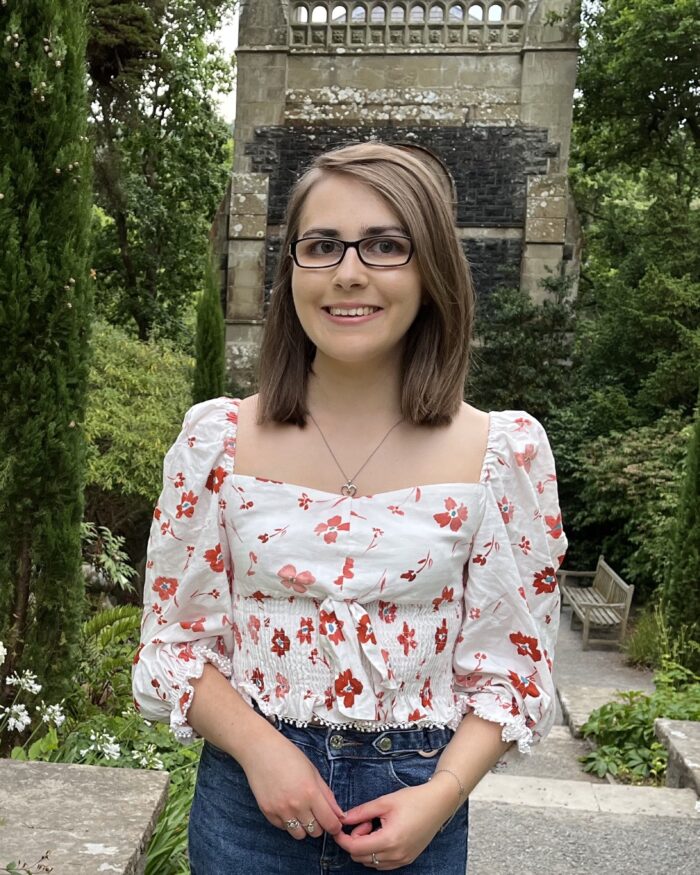 Elin standing on top of steps which lead down to a small stone building with wooden doors and windows. The building is flanked by trees. She wears a white and red floral printed top which has slightly puff sleeves which fall to just below the elbow. It's paired with dark blue high waisted jeans, a silver heart shaped necklace and silver rings. Her shoulder length brown hair is worn straight and she's smiling at the camera.