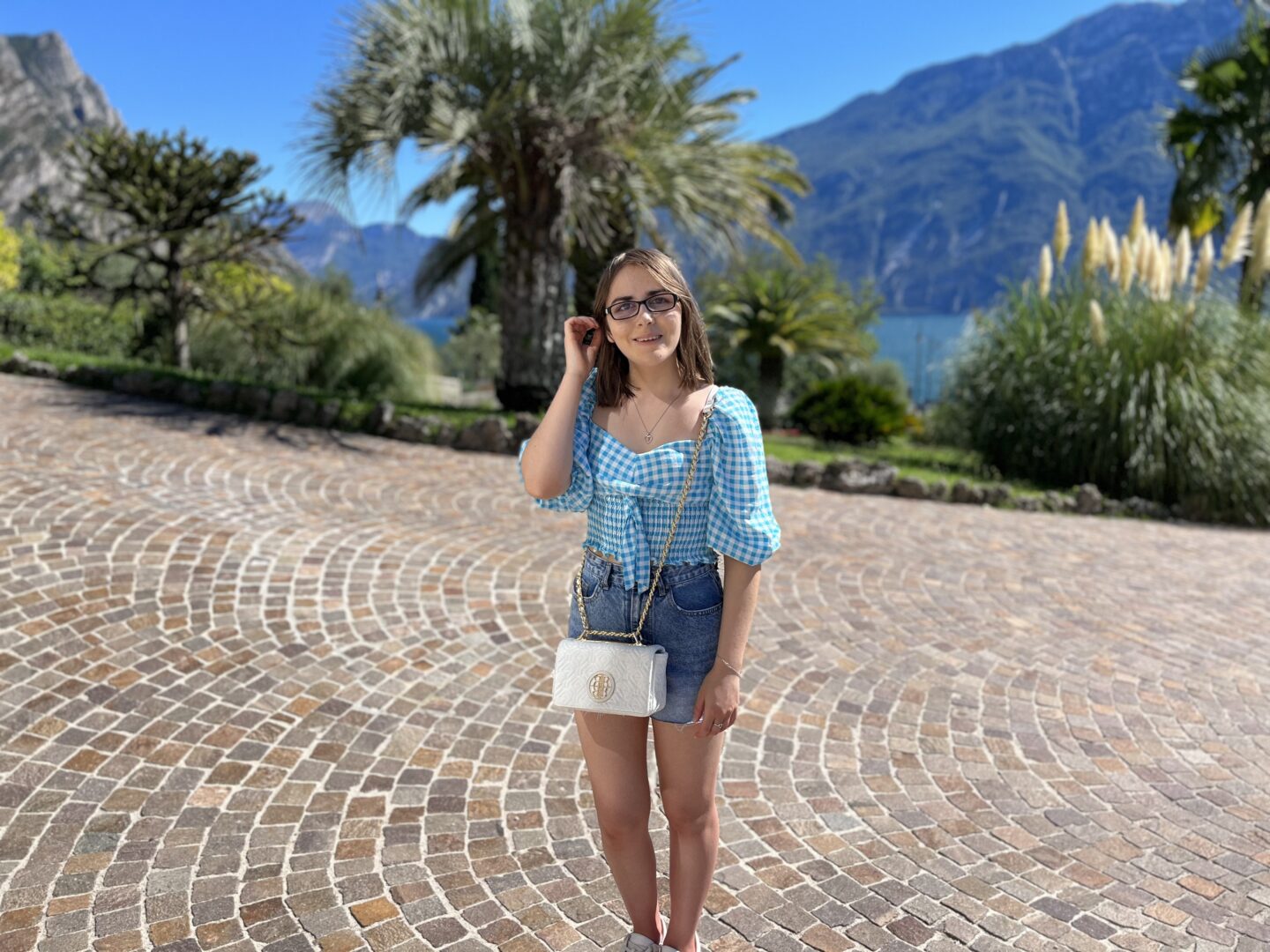 Elin standing on a cobbled path which is framed by trees and plants. Lake Garda peeks out from the trees, and mountains dominate the background. Elin wears a white and blue gingham crop top which has puff sleeves which fall to just beneath her elbow. It's paired with denim shorts, a white handbag with gold hardware and white trainers. She pushes a strand of hair away from her face with her right hand and is smiling at the camera.