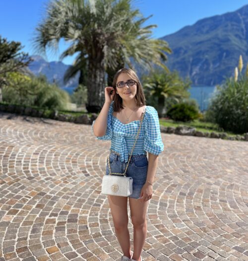 Elin standing on a cobbled path which is framed by trees and plants. Lake Garda peeks out from the trees, and mountains dominate the background. Elin wears a white and blue gingham crop top which has puff sleeves which fall to just beneath her elbow. It's paired with denim shorts, a white handbag with gold hardware and white trainers. She pushes a strand of hair away from her face with her right hand and is smiling at the camera.