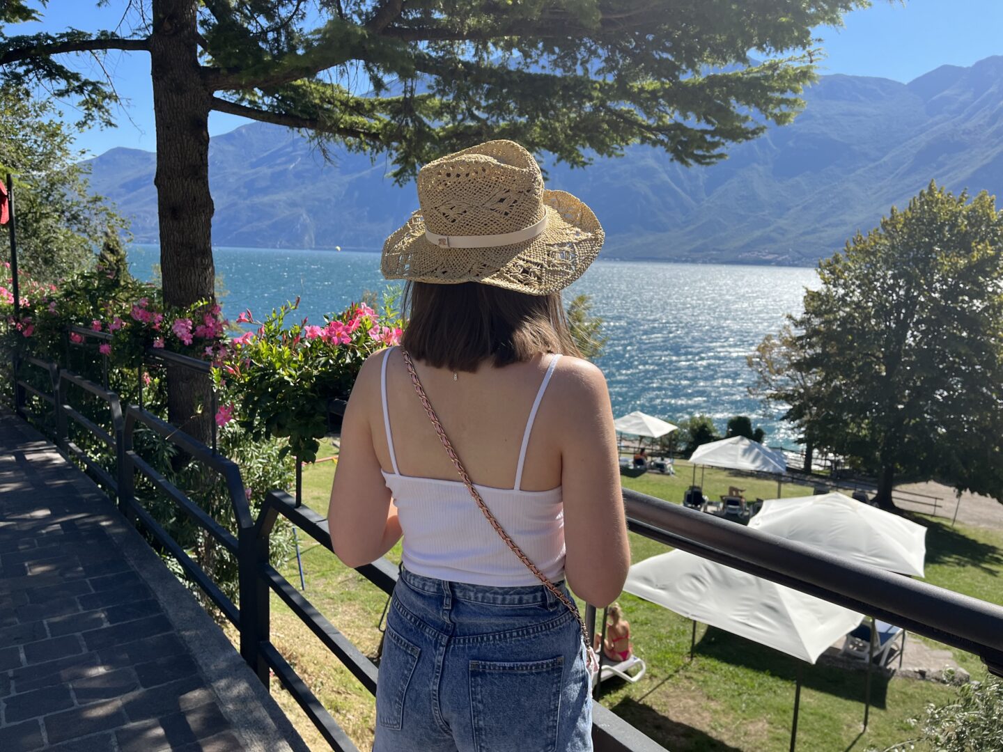 Elin looking at a view of a lake which is framed by mountains. The sun shines on the water. She standing by a black railing which is adorned with pretty pink flowers.
