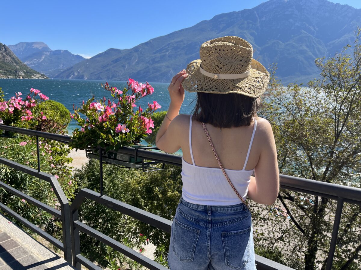 Elin standing with her back to the camera. She is looking out over a lake which is framed by mountains. It's a sunny day and the sky is bright blue. Her left hand holds the edge of a straw hat on her head and her shoulder length brown hair is fluttering in the wind. She wears a white ruched cami top with denim high waisted shorts.