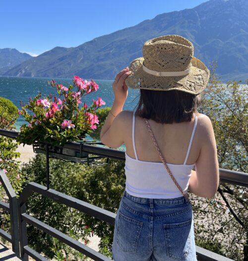 Elin standing with her back to the camera. She is looking out over a lake which is framed by mountains. It's a sunny day and the sky is bright blue. Her left hand holds the edge of a straw hat on her head and her shoulder length brown hair is fluttering in the wind. She wears a white ruched cami top with denim high waisted shorts.