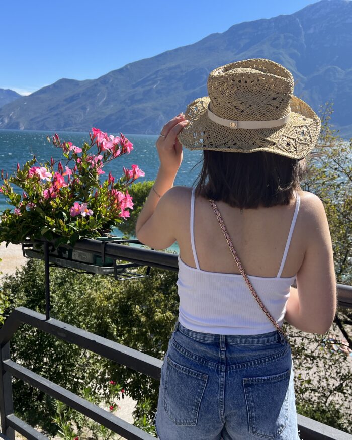 Elin standing with her back to the camera. She is looking out over a lake which is framed by mountains. It's a sunny day and the sky is bright blue. Her left hand holds the edge of a straw hat on her head and her shoulder length brown hair is fluttering in the wind. She wears a white ruched cami top with denim high waisted shorts.