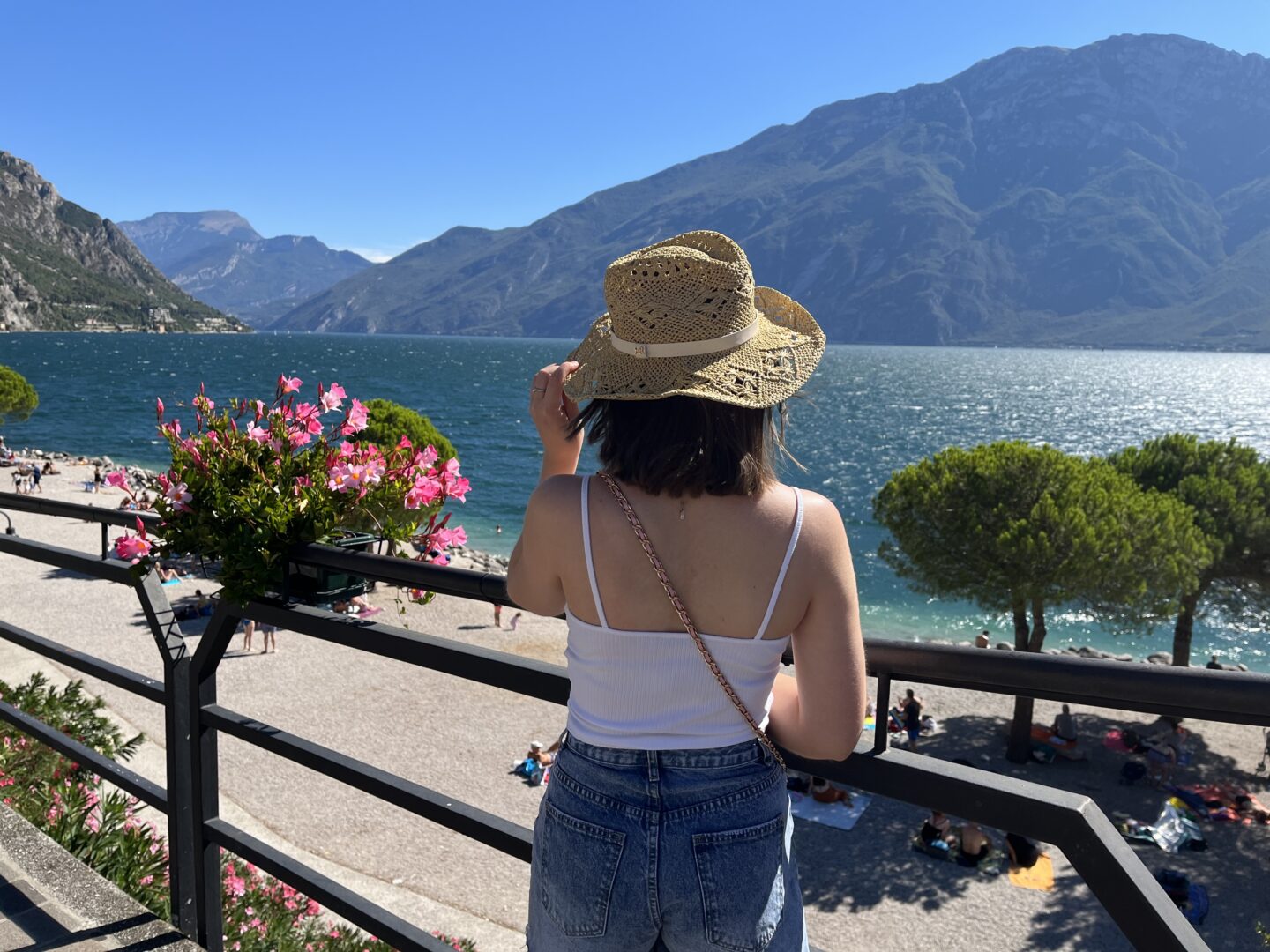 Elin standing with her back to the camera. She is looking out over a lake which is framed by mountains. It's a sunny day and the sky is bright blue. Her left hand holds the edge of a straw hat on her head and her shoulder length brown hair is fluttering in the wind. She wears a white ruched cami top with denim high waisted shorts.