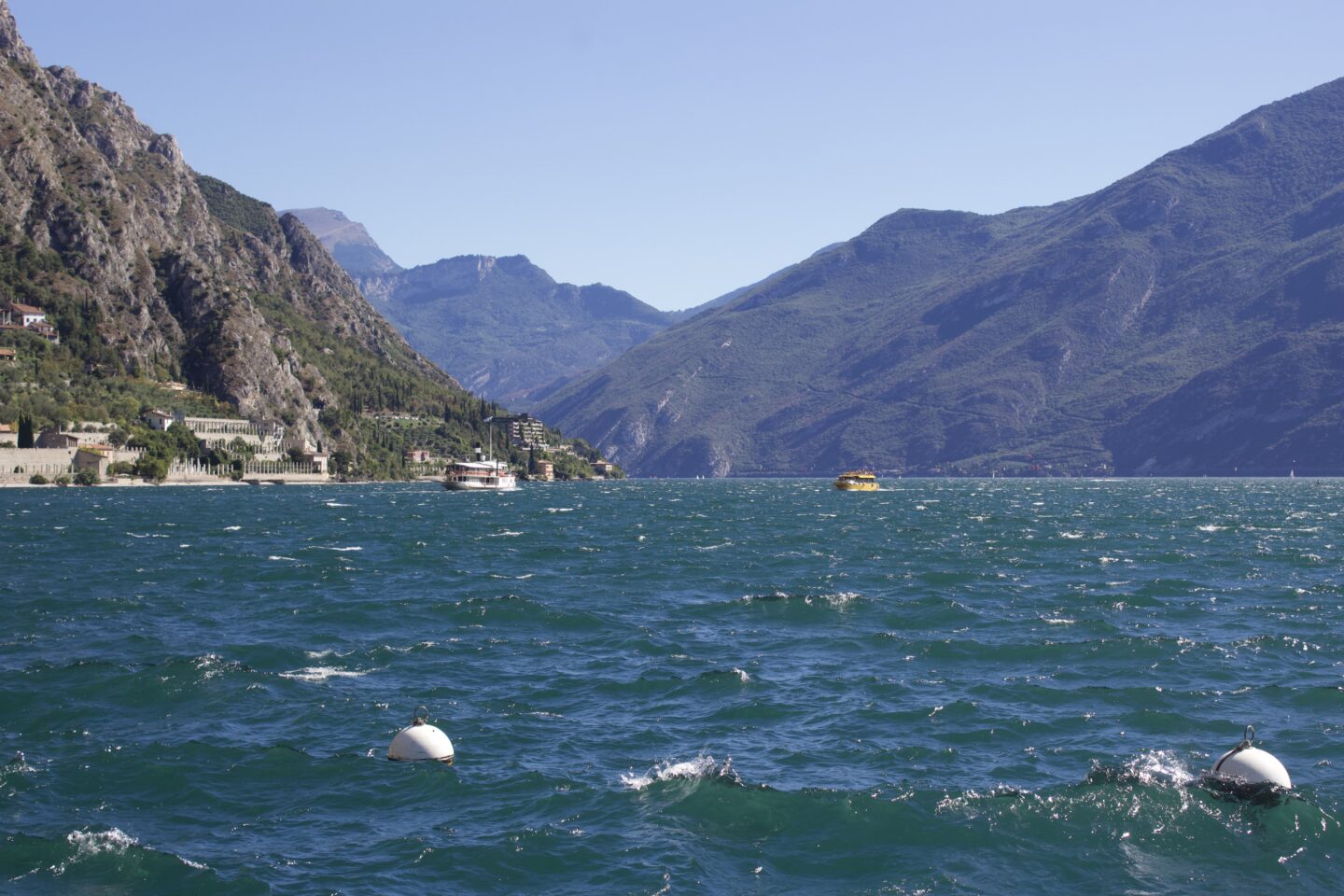 Sun shines on a lake which is framed by a cliffside on one side and mountains on the other. Italian houses line the bottom of the cliff and boat buoys float in the water.
