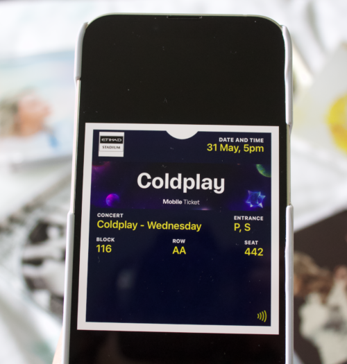 A Coldplay concert ticket showing on a phone screen. The details of the gig, including date and seat number is in white writing on a black background. The phone is held above a collection of CD's which are laid out on a bed, all are blurred in the background.
