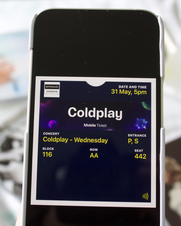 A Coldplay concert ticket showing on a phone screen. The details of the gig, including date and seat number is in white writing on a black background. The phone is held above a collection of CD's which are laid out on a bed, all are blurred in the background.