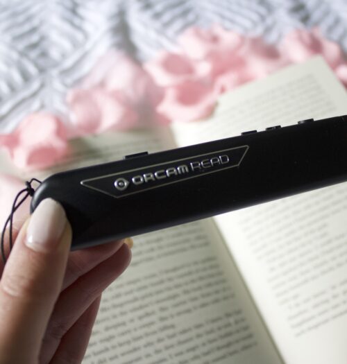 Elin's hand holding the OrCam Read, a black slim pen-style device. It's held over an open book which is placed on a white waffle style duvet and is surrounded by fake pink flower petals. The book and flowers are blurred. Elin's nails are painted a baby pink colour.