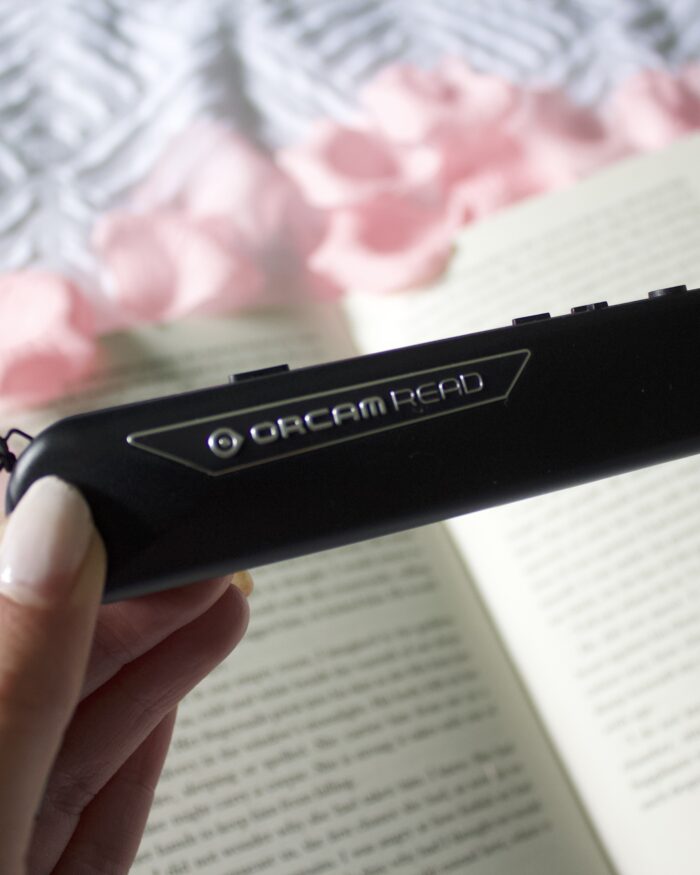 Elin's hand holding the OrCam Read, a black slim pen-style device. It's held over an open book which is placed on a white waffle style duvet and is surrounded by fake pink flower petals. The book and flowers are blurred. Elin's nails are painted a baby pink colour.