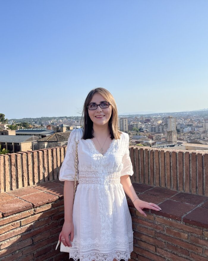 Elin standing with her hand on a stone wall which overlooks the city of Girona in Spain. She wears a white mini skater dress which has lace detailing around the waist, down the centre, along the hem and on the sleeves which fall just below her elbows. It’s paired with a white handbag which has gold hardware. Her shoulder length brown hair is worn straight and she’s smiling broadly at the camera.
