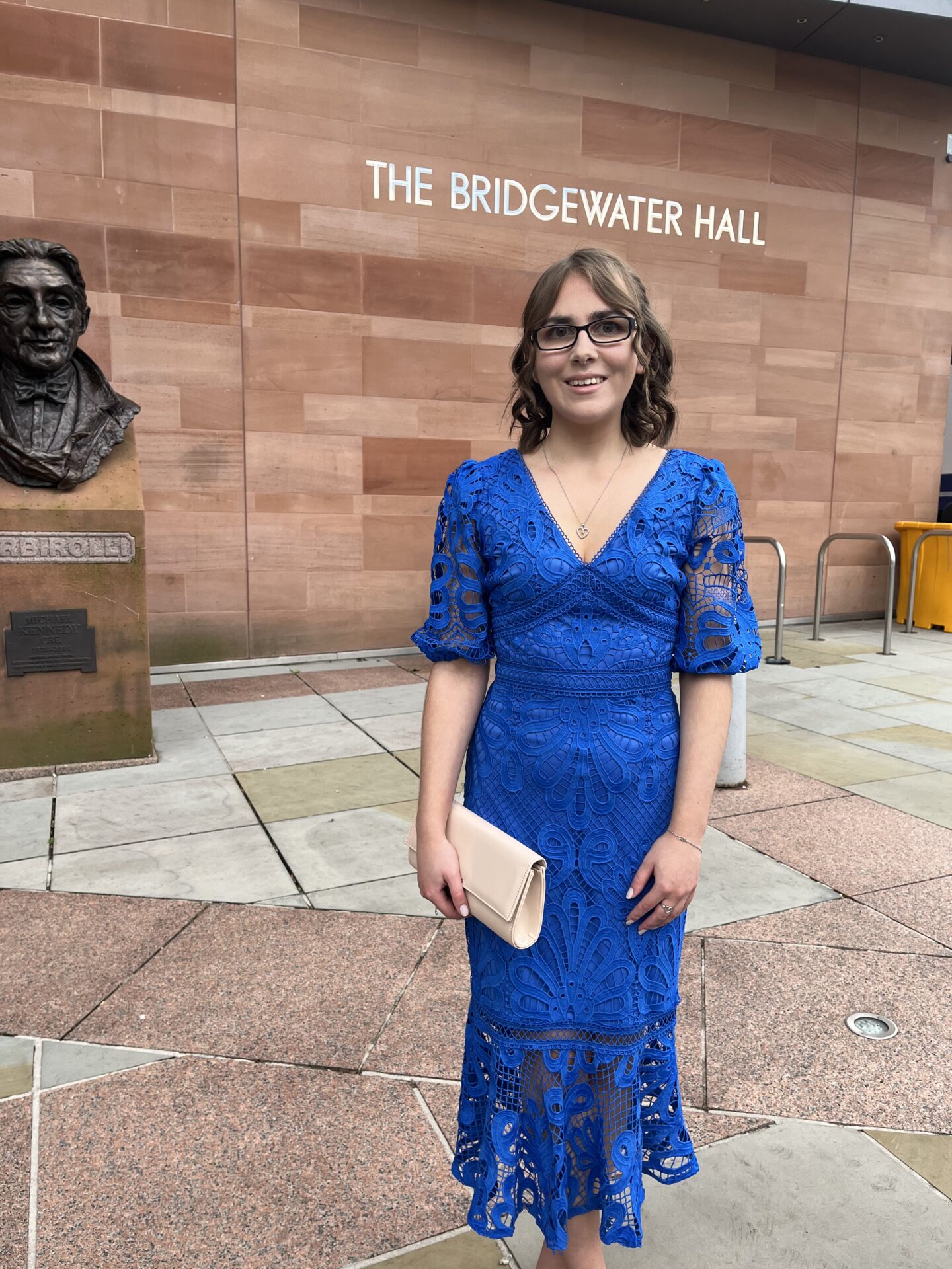 Elin standing in front of The Bridgewater Hall which has the venue's name up in bold white capital letters on the side of it. She wears an all over lace blue midi dress which has a v-neckline and lace see through sleeves that fall to just above the elbow. A see through lace insert also falls from just above the knee to the bottom of the dress. It's paired with a nude clutch bag which Elin holds in her right hand and matching nude heels. Her brown hair is curled and styled into a braided half-up, half-down do. She's smiling widely at the camera.