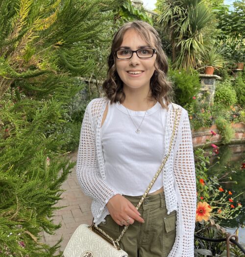 Elin standing in a lush garden, smiling broadly at the camera. She has shoulder length brown hair which is styled into a curly half-up, half-down do and wears black framed glasses. Her outfit is a white tank top with a white crochet cardigan over it, paired with khaki cargo shorts. It's accessorised with a delicate heart shaped necklace and a white cross body bag which has gold hardware. Behind her is a small pond surrounded by various plants and flowers with a reflection of the sky and greenery on the water's surface.