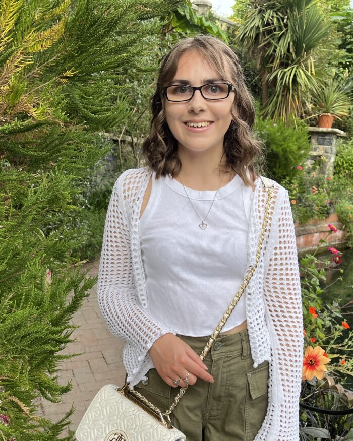 Elin standing in a lush garden, smiling broadly at the camera. She has shoulder length brown hair which is styled into a curly half-up, half-down do and wears black framed glasses. Her outfit is a white tank top with a white crochet cardigan over it, paired with khaki cargo shorts. It's accessorised with a delicate heart shaped necklace and a white cross body bag which has gold hardware. Behind her is a small pond surrounded by various plants and flowers with a reflection of the sky and greenery on the water's surface.