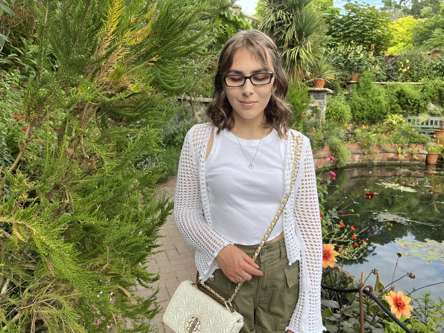 Elin looking down reflectively. She has shoulder length brown hair which is styled into a curly half-up, half-down do and wears black framed glasses. Her outfit is a white tank top with a white crochet cardigan over it, paired with khaki cargo shorts. It's accessorised with a delicate heart shaped necklace and a white cross body bag which has gold hardware. Behind her is a small pond surrounded by various plants and flowers with a reflection of the sky and greenery on the water's surface.