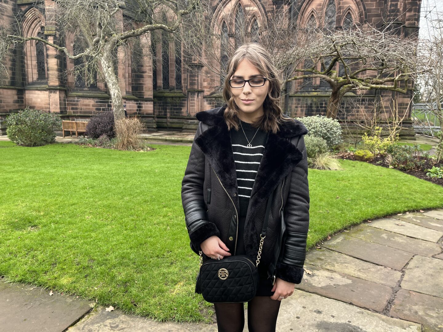 Elin standing on a stone path in front of a large cathedral which is bordered by a manicured lawn. She's looking down reflectively.