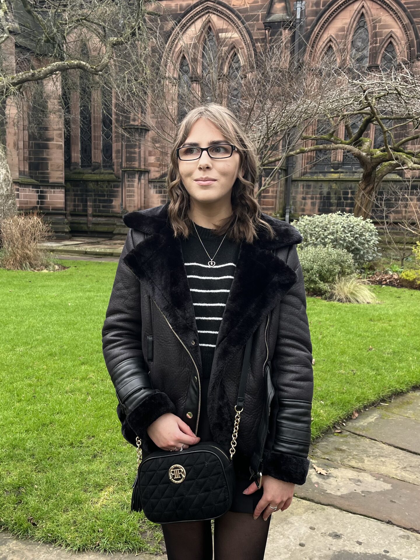 Elin standing on a stone path in front of a large cathedral which has large gothic style windows. The path is bordered by a manicured lawn. She wears a black faux fur lined aviator jacket over a black and white crop jumper and black skort. The outfit is accessorised with a black crossbody bag which has gold hardware, silver jewellery and Elin's trademark black glasses which have diamonds on the side.. Her hair is styled into curls and is worn in a half-up, half-down do. She's looking away reflectively to her right.