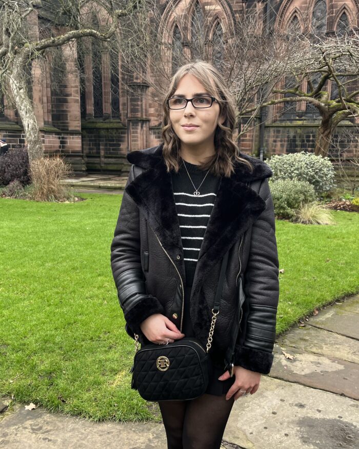 Elin standing on a stone path in front of a large cathedral which has large gothic style windows. The path is bordered by a manicured lawn. She wears a black faux fur lined aviator jacket over a black and white crop jumper and black skort. The outfit is accessorised with a black crossbody bag which has gold hardware, silver jewellery and Elin's trademark black glasses which have diamonds on the side.. Her hair is styled into curls and is worn in a half-up, half-down do. She's looking away reflectively to her right.