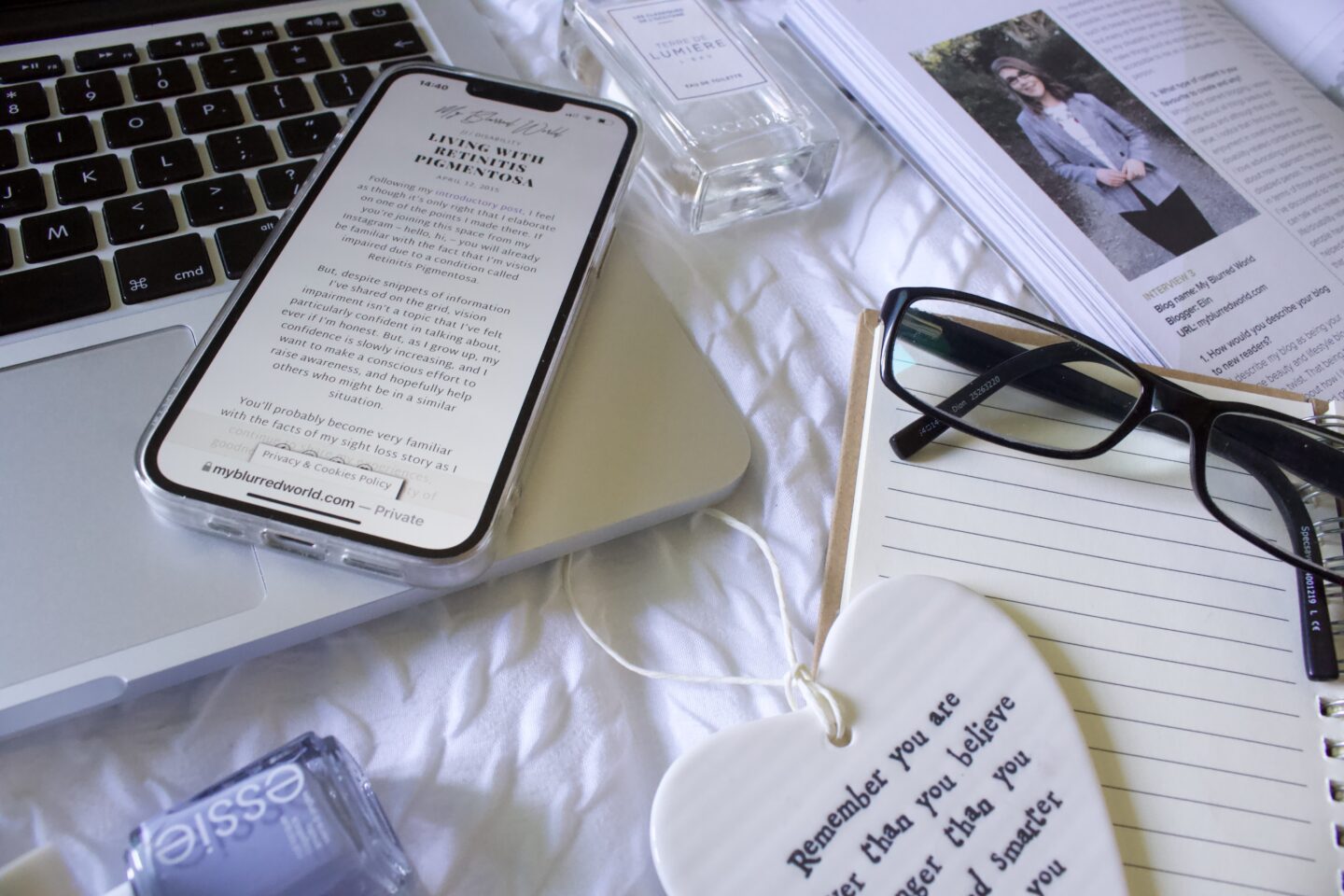 Elin's second published blog post titled 'Living with Retinitis Pigmentosa' displayed an a phone screen. The phone rests on the corner of a laptop keyboard. Beside it is an open notebook with a pair of black framed glasses resting on it. The notebook overlaps with an open magazine which shows a photo of Elin from 2018. There's bottle of blue nail polish in the bottom left corner and a perfume bottle between the laptop and the magazine.