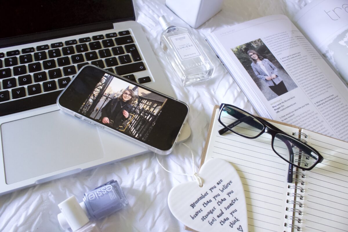A phone displaying a photo of a younger Elin in a black top and leather jacket rests on the edge of a laptop keyboard. It's surrounded by a perfume bottle, two nail polishes, a copy of Blogosphere magazine which is open to Elin's #InTheSpotlight feature from 2018 and an open notebook with a pair of black framed glasses resting on it along with a heart shaped hanging ornament with words that read 'Remember that you are braver than you believe, stronger than you feel and smarter than you think.
