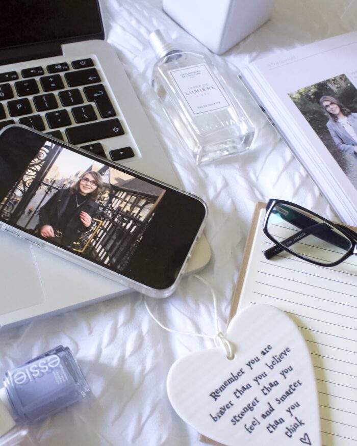 A phone displaying a photo of a younger Elin in a black top and leather jacket rests on the edge of a laptop keyboard. It's surrounded by a perfume bottle, two nail polishes, a copy of Blogosphere magazine which is open to Elin's #InTheSpotlight feature from 2018 and an open notebook with a pair of black framed glasses resting on it along with a heart shaped hanging ornament with words that read 'Remember that you are braver than you believe, stronger than you feel and smarter than you think.