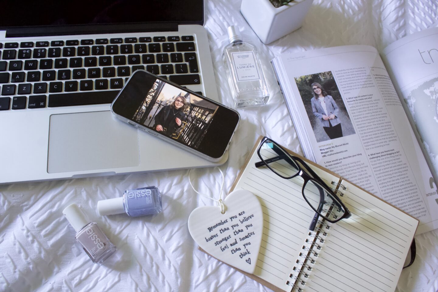 A phone displaying a photo of a younger Elin in a black top and leather jacket rests on the edge of a laptop keyboard. It's surrounded by a perfume bottle, two nail polishes, a copy of Blogosphere magazine which is open to Elin's #InTheSpotlight feature from 2018 and an open notebook with a pair of black framed glasses resting on it along with a heart shaped hanging ornament with words that read 'Remember that you are brave than you believe, stronger than you feel and smarter than you think.