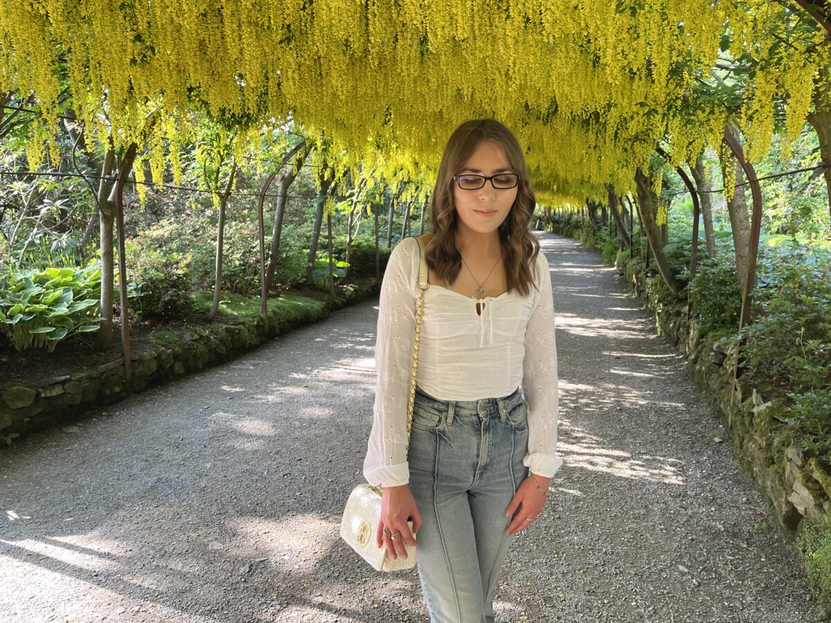 Elin standing on a pathway in a garden, she's looking down reflectively. She has long, wavy brown hair and is wearing black framed glasses, a white, long-sleeved ruched top which has a square neckline, high-waisted light blue jeans and a white handbag with gold hardware. The pathway is shaded by an overhead canopy of bright yellow flowers hanging down from the trees, creating a tunnel-like effect.
