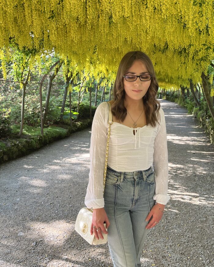 Elin standing on a pathway in a garden, she's looking down reflectively. She has long, wavy brown hair and is wearing black framed glasses, a white, long-sleeved ruched top which has a square neckline, high-waisted light blue jeans and a white handbag with gold hardware. The pathway is shaded by an overhead canopy of bright yellow flowers hanging down from the trees, creating a tunnel-like effect.