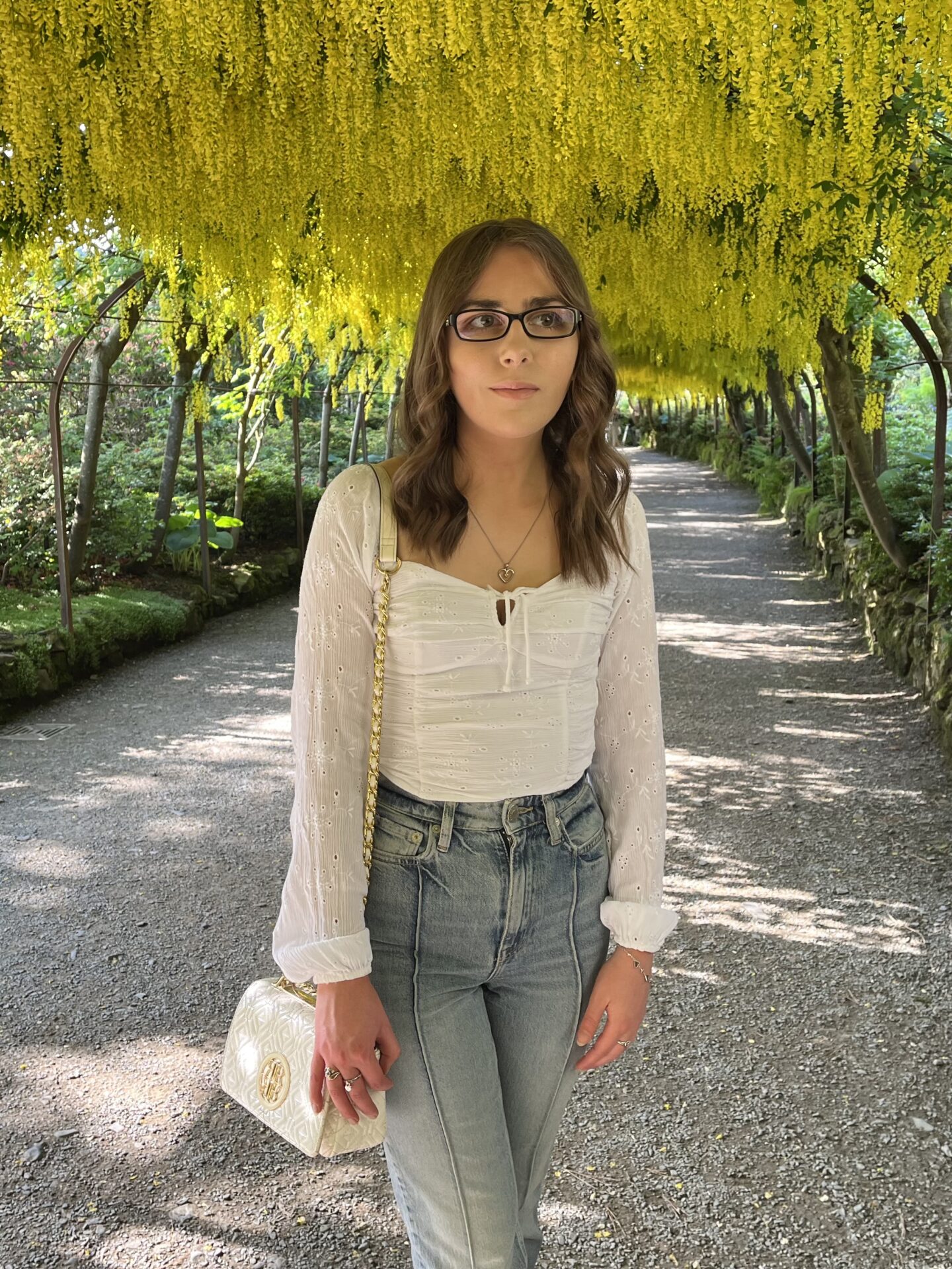 Elin standing on a pathway in a garden, she's looking away to her left. She has long, wavy brown hair and is wearing black framed glasses, a white, long-sleeved ruched top, high-waisted light blue jeans and a white handbag. The pathway is shaded by an overhead canopy of bright yellow flowers hanging down from the trees, creating a tunnel-like effect.