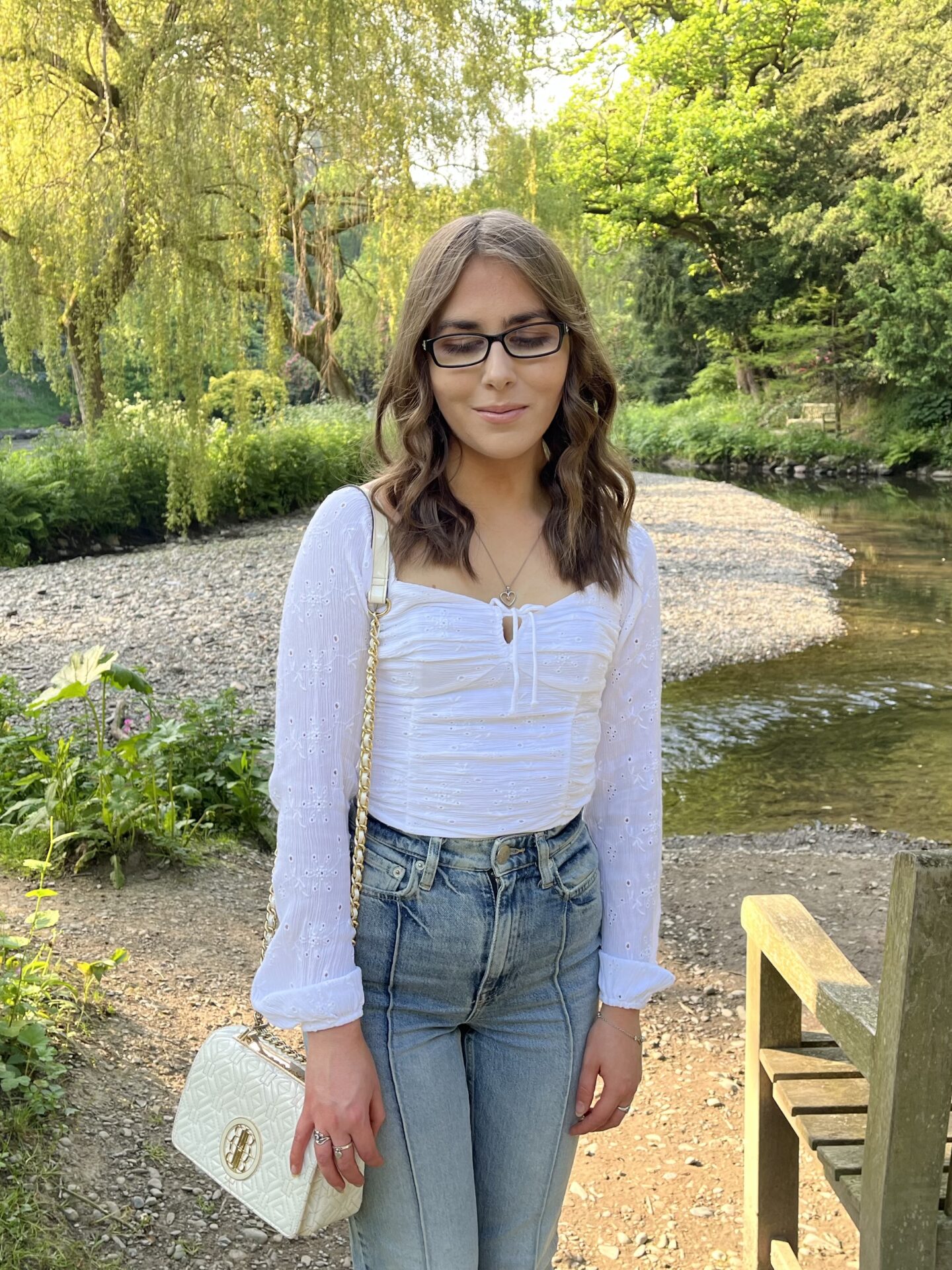 Elin standing by a wooden bench in front of a stream which is surrounded by lush greenery and trees. She's looking down reflectively. She is dressed in a white long sleeve ruched top which has a square neckline, light blue high waisted jeans and a white handbag with a gold chain strap. The sun shines on the trees in the background.