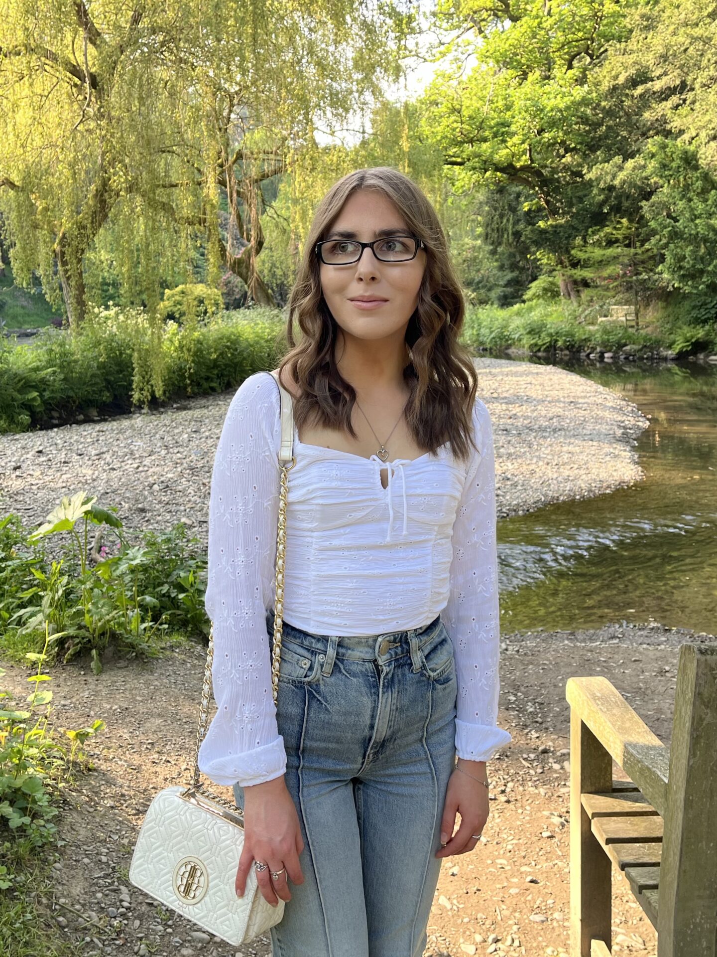 Elin standing by a wooden bench in front of a stream which is surrounded by lush greenery and trees. She has long wavy brown hair and is looking away to her right. She is dressed in a white long sleeve ruched top which has a square neckline, light blue high waisted jeans and a white handbag with a gold chain strap. The sun shines on the trees in the background.