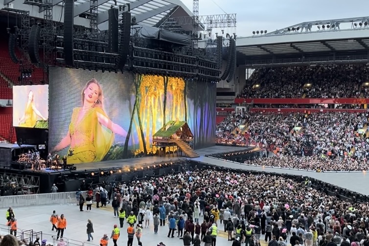 Another close up of Taylor on the large screen during the Folklore/Evermore sister set. Her dress looks yellow because of the lighting and her head lulls slightly to the side as she closes her eyes. The stage setup includes vibrant, colourful decorations, including a backdrop of trees/foliage. The photo is taken from the left side of the stage which is long and juts out into the crowd who are standing around it. Part of the stand on the far side is visible too.