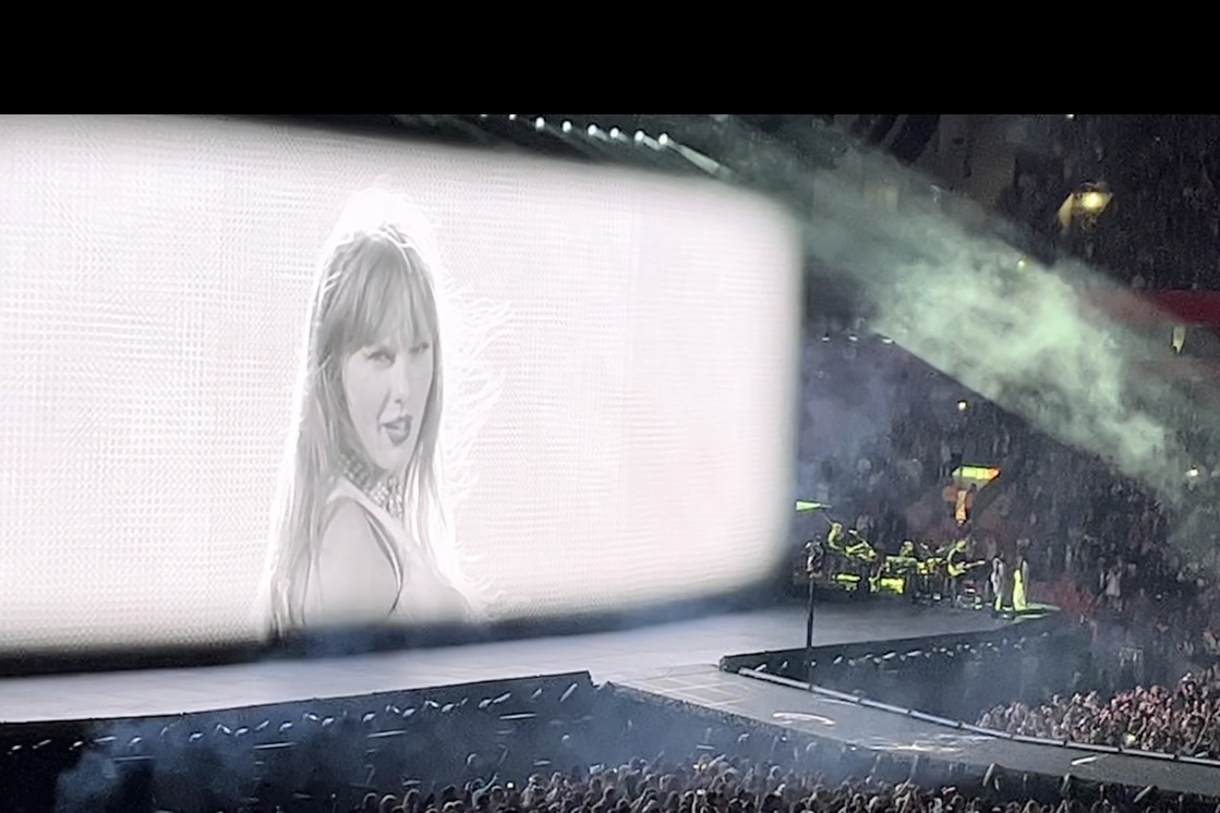 The big screen shows a black and white close-up of Taylor during her performance of Who’s Afraid of Little Old Me? She has long hair with a fringe and is looking over her shoulder, with a confident, menacing expression. The stage is surrounded by the crowd, and there are some musicians and performers visible on the right side of the stage. Smoke and lighting add to the dramatic feel of the song.