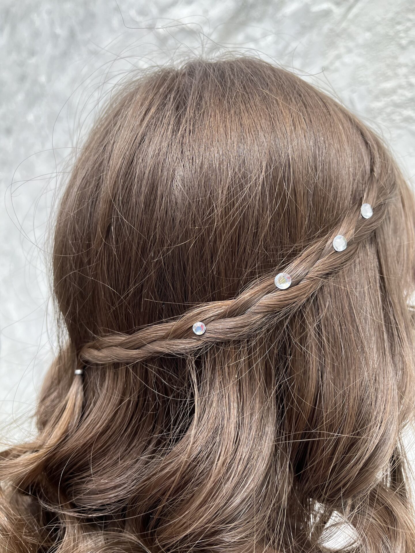 Elin's hairstyle taken from a different angle. Her brown locks are styled into loose curls with two small braids starting from each side and meeting at the back. The braids are adorned with four small circular gems that catch the light.