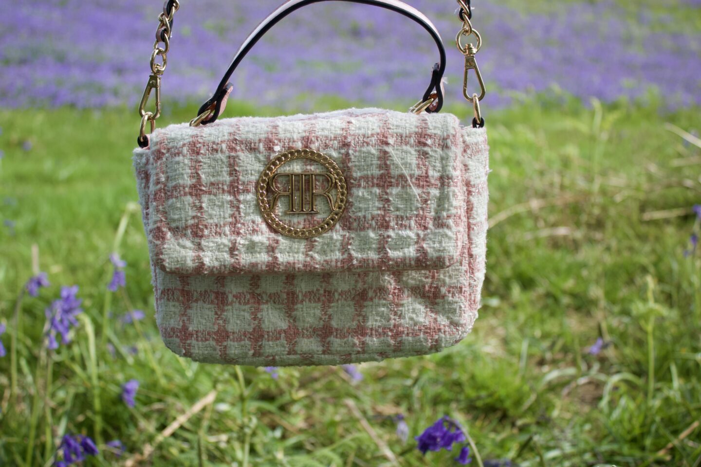 A close up of a pink and white checkered handbag. It has a gold circular emblem in the centre with the letters 'RR' intertwined. It hangs by a chain strap. The background is blurred but shows a field of bright bluebells.
