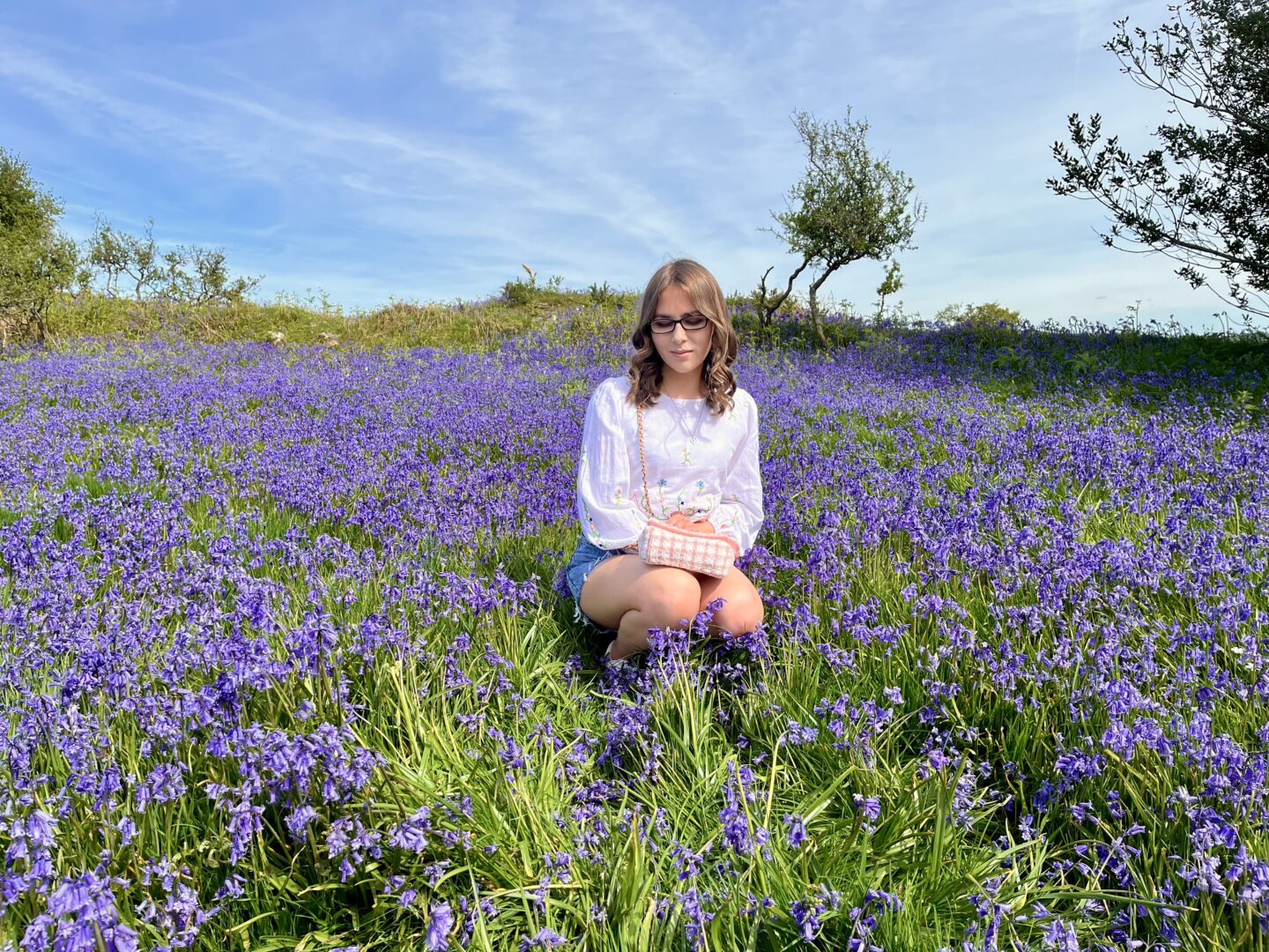Elin crouching down amongst a carpet of brightly coloured bluebells. She's looking down reflectively. She wears a white long sleeve crop top which has floral details on the sleeves and hem, paired with light blue denim shorts, silver jewellery and a white and pink handbag which rests on her lap. Her brown hair is styled into loose curls.