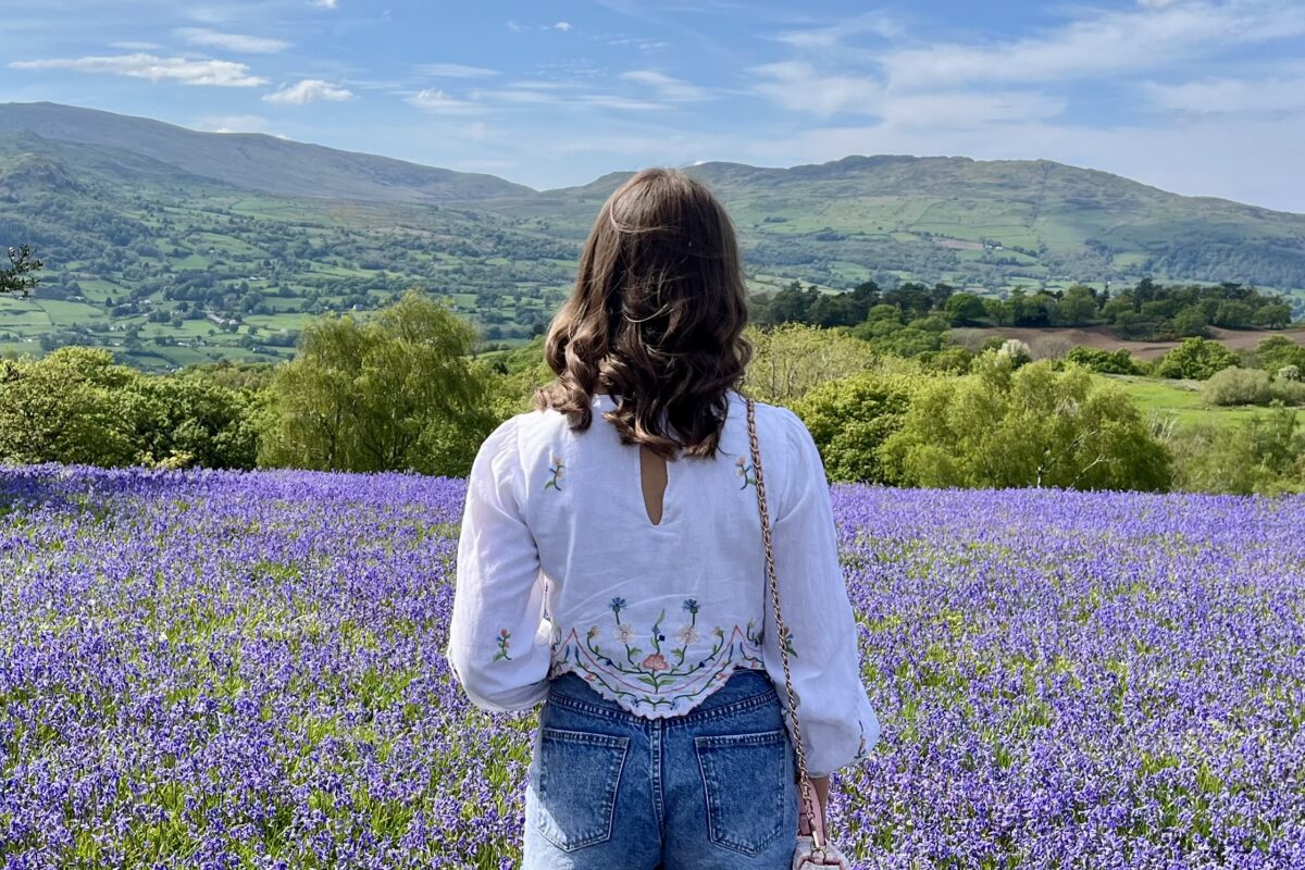 Elin standing in a field carpeted with bluebells. She faces away from the camera, looking out at a view of mountains under a blue sky. She wears a white long-sleeve top with floral details and blue high waisted shorts. A small pink bag hangs from her right shoulder and her brown hair is styled into curls.