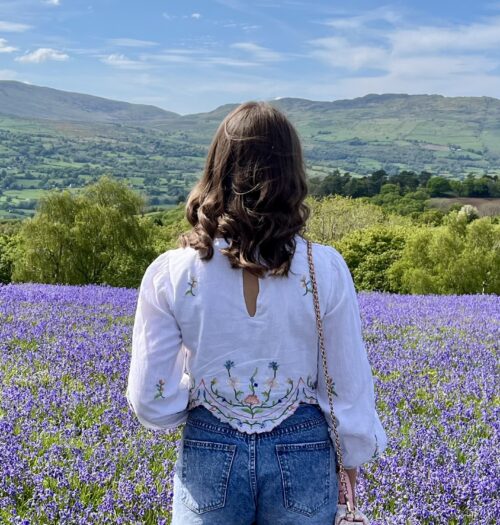 Elin standing in a field carpeted with bluebells. She faces away from the camera, looking out at a view of mountains under a blue sky. She wears a white long-sleeve top with floral details and blue high waisted shorts. A small pink bag hangs from her right shoulder and her brown hair is styled into curls.