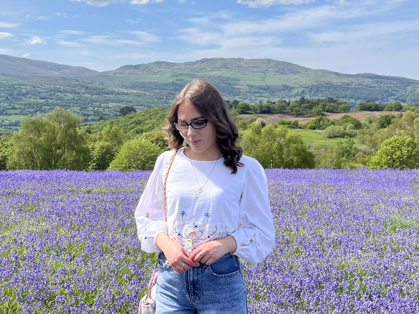 Elin standing in a field full of bluebells with hills and mountains in the background. She wears a white crop top which has floral details on the sleeves and hem, it's paired with denim high waisted shorts, silver jewellery and a small light pink boucle handbag which hangs from her right shoulder. Elin is looking down reflectively.