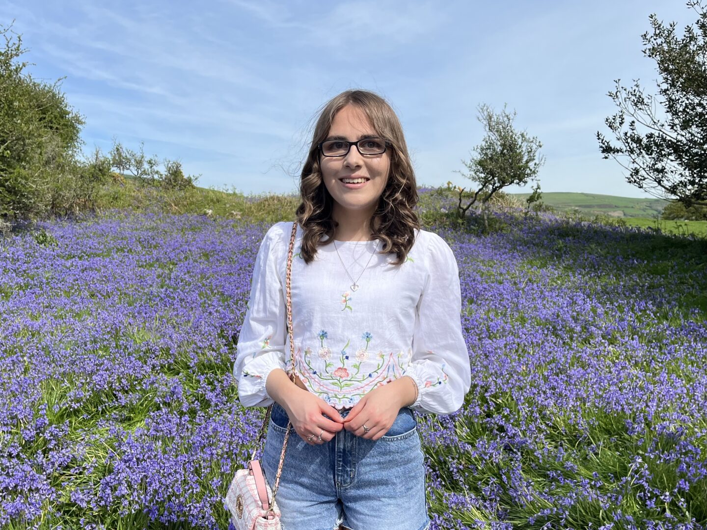 Elin standing in a field full of bluebells. She wears a white crop top which has floral details on the sleeves and hem, it's paired with denim high waisted shorts, silver jewellery and a small light pink boucle handbag which hangs from her right shoulder. Her brown hair is styled into loose curls and she's smiling broadly at the camera.