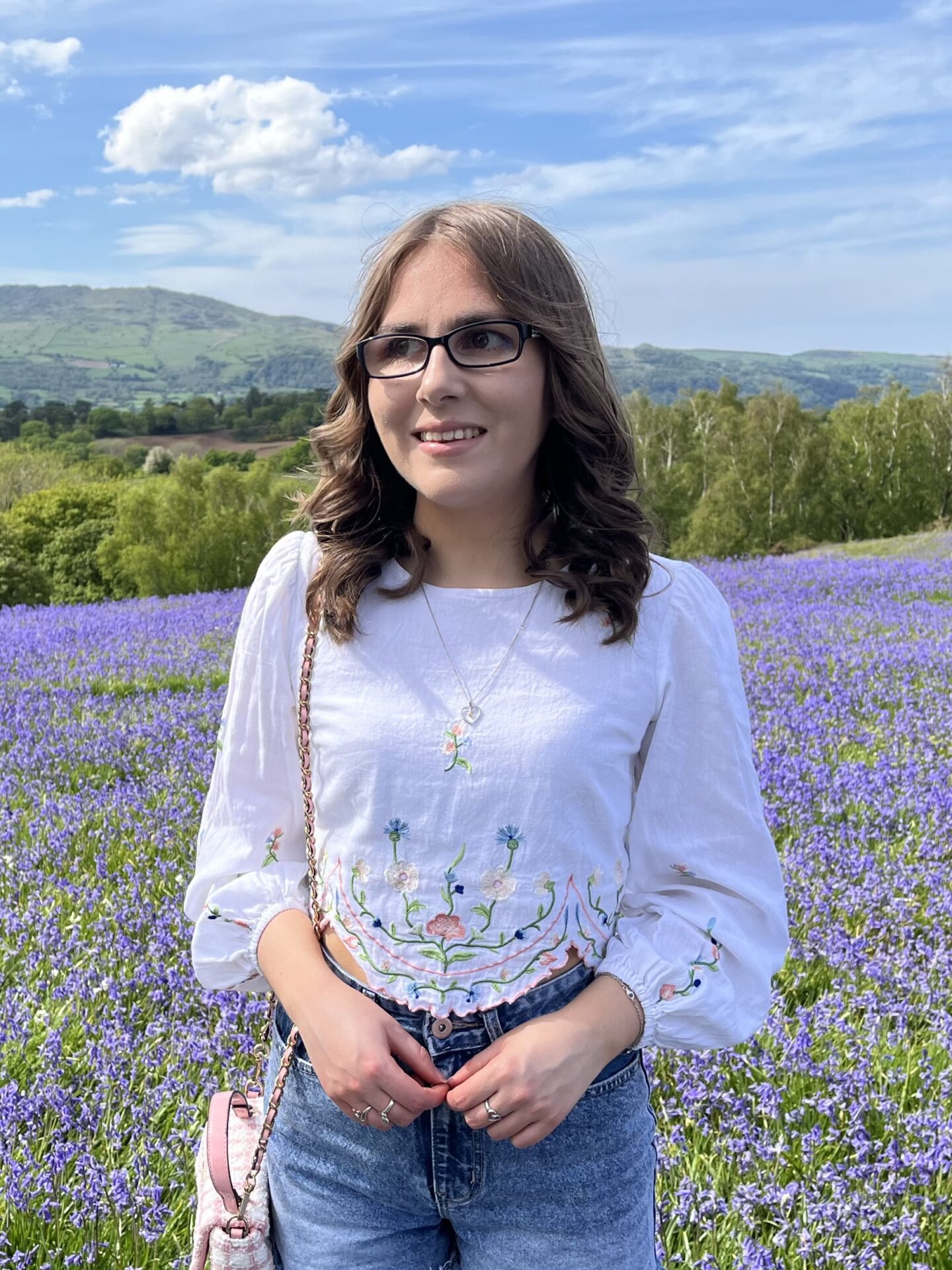 Elin standing in a field full of bluebells with mountains in the distance. She's looking away from the camera to her right, smiling broadly.