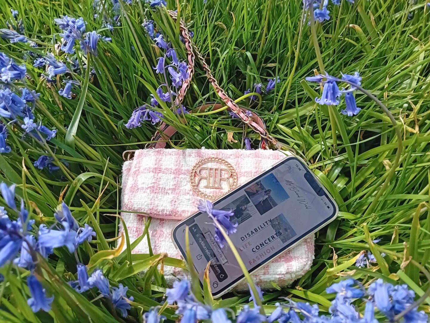 A small pink and white check handbag which has a gold emblem on the front resting amongst brightly coloured bluebells. A phone rests on the edge of the bag displaying the My Blurred World blog homepage.